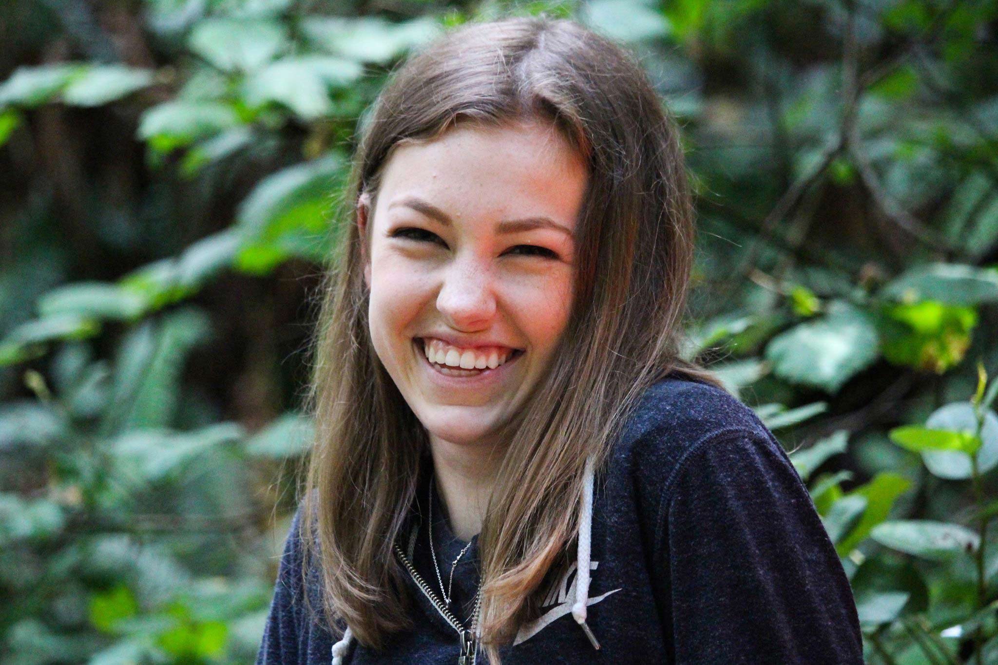 Girl sits in nature surrounded by trees smiling
