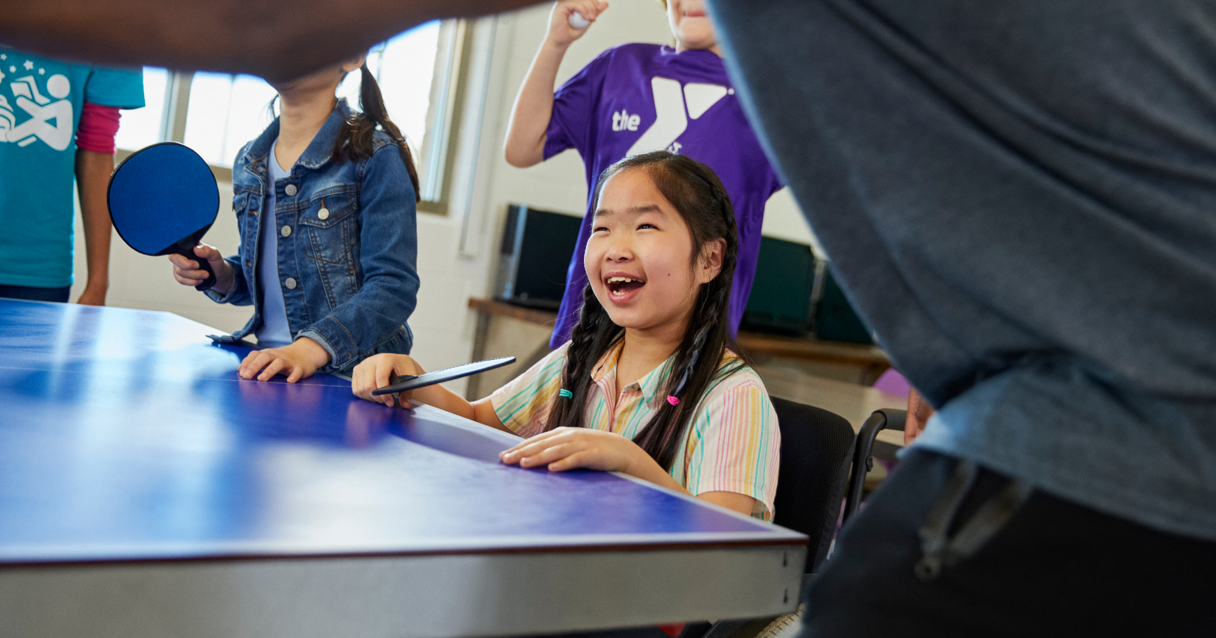 Smiling little girl standing at ping pong table with a ping pong paddle in hand with other people around her also playing ping pong